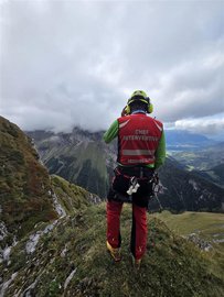 Un secouriste de la colonne de secours se tient sur une crête de montagne, dos à la caméra, et observe une vallée. Il porte des vêtements d'intervention avec l'inscription « CHEF INTERVENTION SECOURS ALPIN » sur sa veste rouge.
