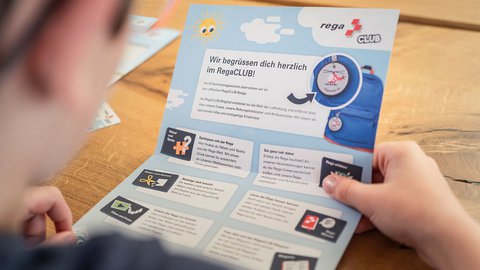Ein Kind hält ein Willkommens-Leporello des Rega-Clubs in den Händen und liest die Informationen.