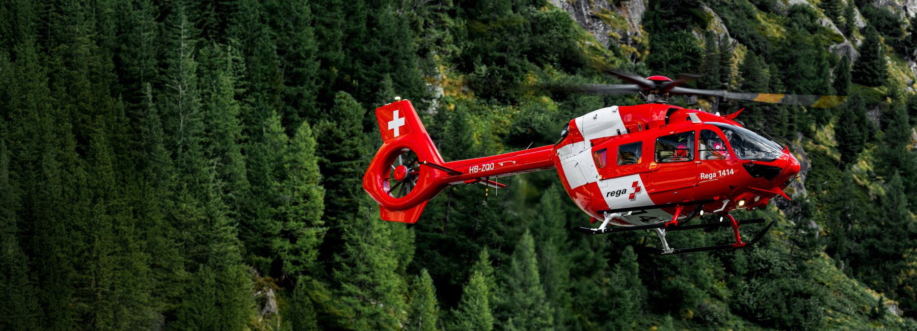 A Rega helicopter flies in front of a wooded mountain.