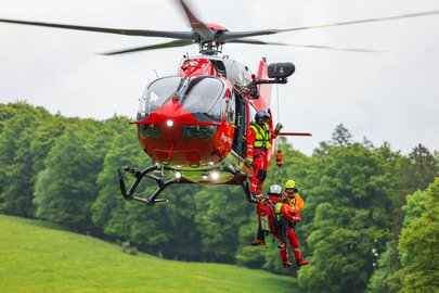 Ein roter Rega-Helikopter schwebt in der Luft, während ein Rettungsspezialist und eine Person an einer Seilwinde unter dem Helikopter hängen. Im Hintergrund ist eine grüne, bewaldete Hügellandschaft unter einem bewölkten Himmel zu sehen.