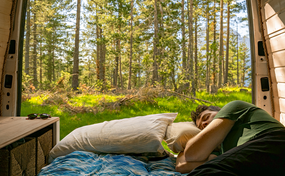 Un homme dort sur un lit dans une camionnette, dont les portes arrière ouvertes offrent une vue sur une forêt ensoleillée.