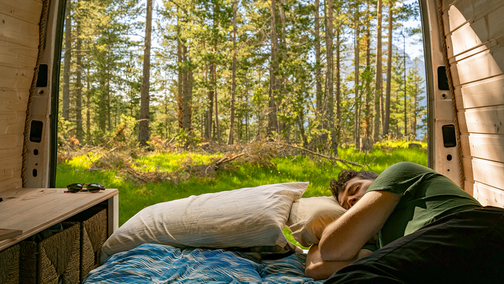 Un uomo dorme su un letto in un furgone, le cui porte posteriori aperte offrono una vista su una foresta soleggiata.