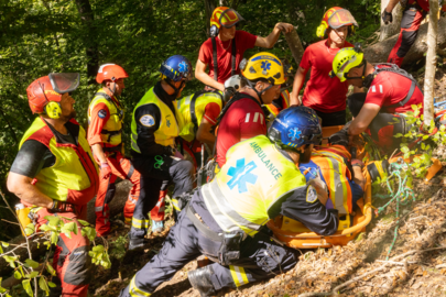 Plusieurs personnes travaillent ensemble dans une forêt pour transporter une personne sur une civière à travers un terrain escarpé. Les secouristes portent des vêtements d'intervention rouges et jaunes et des casques.