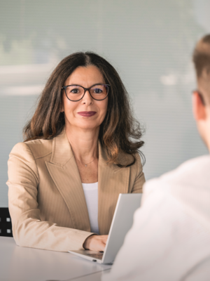 A professionally dressed woman wearing glasses and a beige blazer is sitting at a table. She is looking directly at the camera, while a young man in a white hoodie is sitting in the foreground with his back to the camera. An open laptop is on the table.