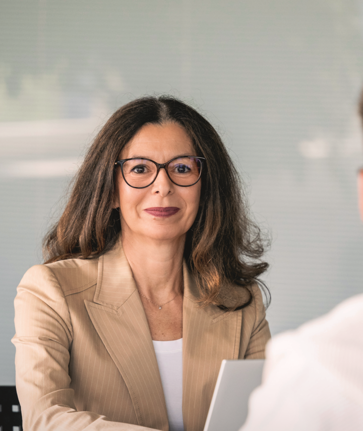 Eine professionell gekleidete Frau, die Brille trägt und einen beigefarbenen Blazer anhat, sitzt an einem Tisch. Sie blickt direkt in die Kamera, während ein junger Mann mit einem weissen Kapuzenpullover im Vordergrund mit dem Rücken zur Kamera sitzt. Auf dem Tisch steht ein offener Laptop.