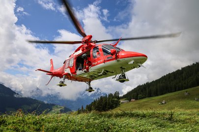 Un hélicoptère de sauvetage AgustaWestland Da Vinci de la Rega est en vol stationnaire à basse altitude au-dessus d'une prairie de montagne. Un spécialiste en sauvetage de la Rega se tient dans la porte latérale ouverte et regarde vers le bas. Un paysage de montagne boisé est visible en arrière-plan.