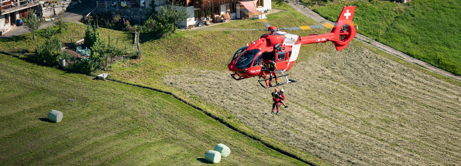Un elicottero della Rega in volo stazionario sopra un prato. Un soccorritore si cala con un verricello per recuperare un paziente.
