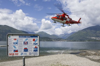 Un elicottero rosso della Rega vola sopra il Lago Maggiore con un paesaggio montano sullo sfondo. In primo piano, un cartello informativo mostra le regole per i bagnanti e i numeri di emergenza (SOS 117).