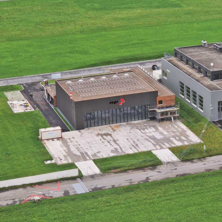 Luftaufnahme der fast fertiggestellten Rega-Basis Mollis und des angrenzenden Hangars, eingebettet in grüne Felder neben der Landebahn.