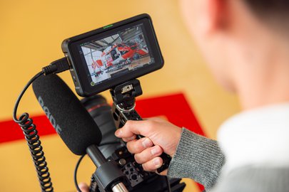 Über die Schulter gefilmt: Ein Mediamatiker-Lehrling hält eine professionelle Kamera mit Mikrofon. Auf dem Kameramonitor ist ein Rega-Helikopter im Hangar zu sehen.