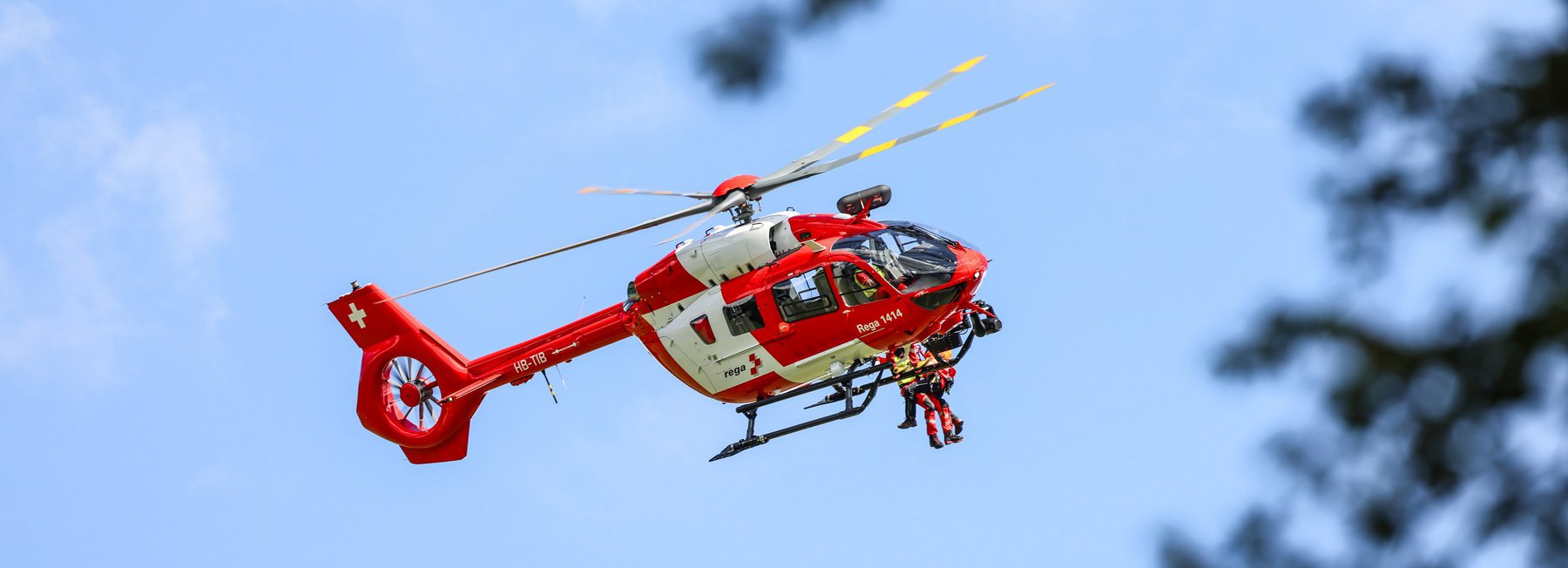 Un elicottero di salvataggio Rega rosso e bianco, un Airbus Helicopters H145, in volo contro un cielo azzurro con nuvole bianche. Un soccorritore è appeso alla corda sotto l'elicottero e sta assicurando una persona ferita in fase di salvataggio. Rami sfocati in primo piano.