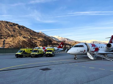 Mehrere Ambulanzfahrzeuge und ein Rega-Ambulanzjet stehen auf dem Vorfeld eines Flughafens in alpiner Umgebung.