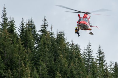 Un hélicoptère de sauvetage AgustaWestland Da Vinci de la Rega transporte un sauveteur par corde au-dessus de la cime d'une forêt. L'hélicoptère est rouge et blanc et vole par ciel couvert.