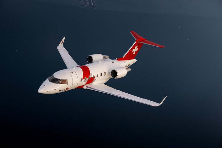 Un jet di soccorso Rega Challenger 650, con la sigla HB-JWA, vola a bassa quota sopra un lago calmo. 