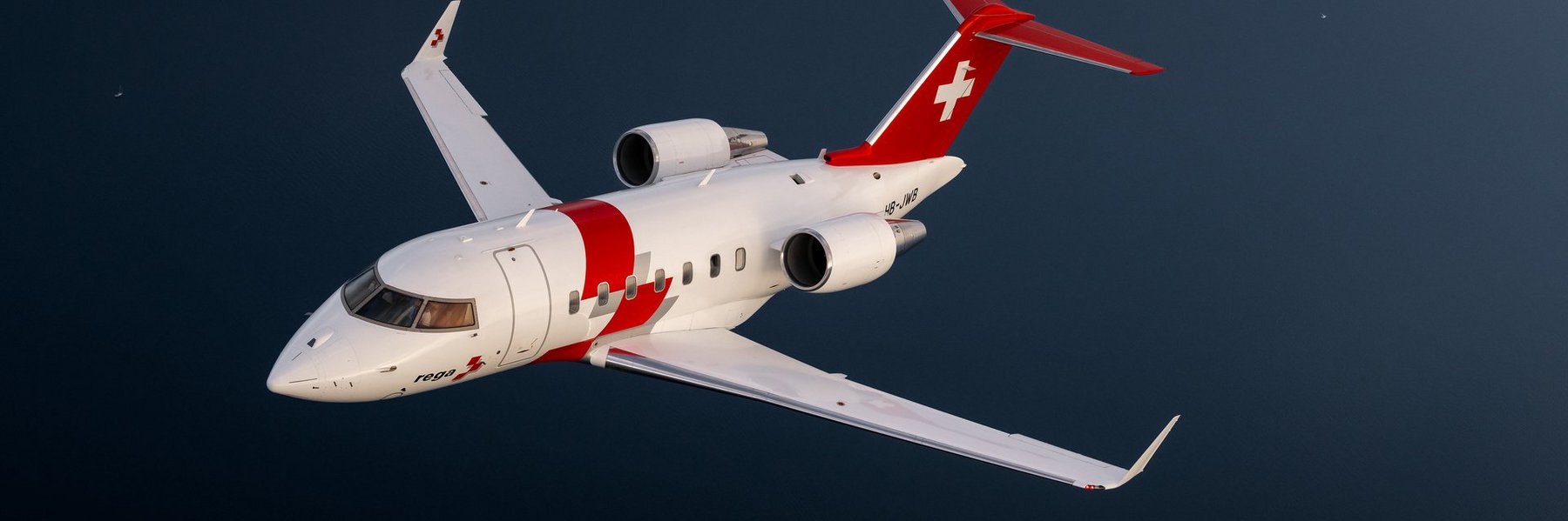 Un jet de sauvetage Rega Challenger 650, immatriculé HB-JWA, vole à basse altitude au-dessus d'un lac calme.