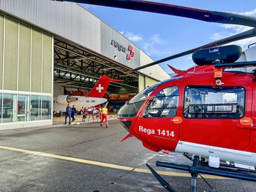 Rega-Rettungshelikopter vor einem Rega-Hangar, während Rettungskräfte einen Patienten zum Ambulanzjet begleiten.