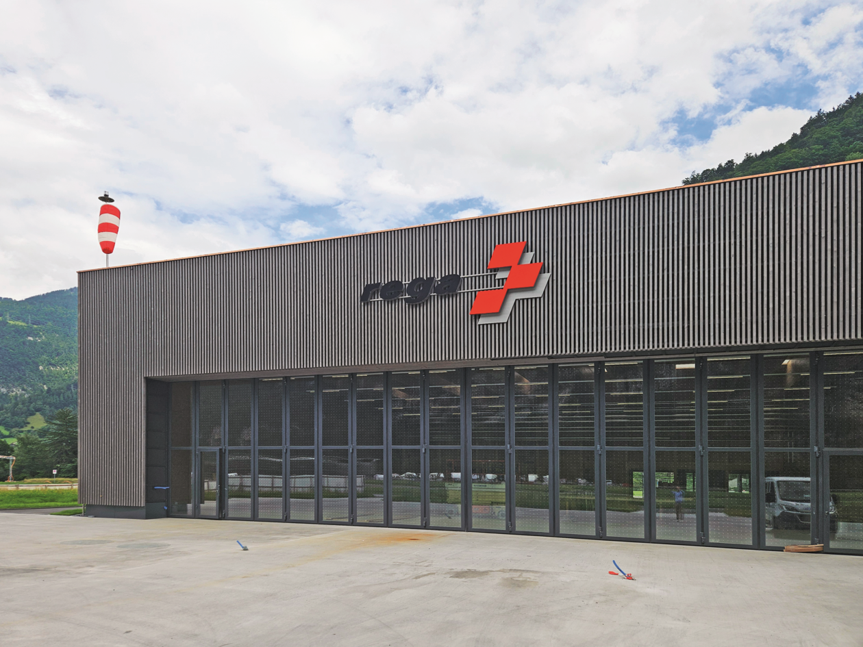 Vorderansicht der neuen, fertiggestellten Rega-Basis Mollis mit Holzlattung, grossen Glastoren und dem Rega-Logo.