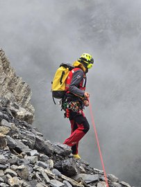 Un secouriste de la colonne de secours se tient sur une pente rocheuse. Il porte des vêtements d'intervention rouges et noirs, un sac à dos jaune et un casque. Il tient une corde rouge dans sa main.