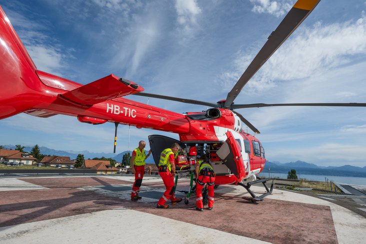 Die Rega-Crew bereitet sich für den Transport in einem roten Rettungshelikopter vor, der auf einem Landeplatz steht. Im Hintergrund sind Häuser und Berge unter einem blauen Himmel mit Wolken zu sehen.