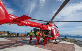 Die Rega-Crew bereitet sich für den Transport in einem roten Rettungshelikopter vor, der auf einem Landeplatz steht. Im Hintergrund sind Häuser und Berge unter einem blauen Himmel mit Wolken zu sehen.