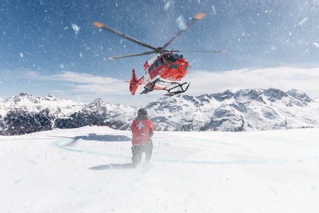 Zum Downloadformular für das Bild Rega-Helikopter im Wintereinsatz auf einer Skipiste mit einem Mitarbeiter des Pistendienstes.