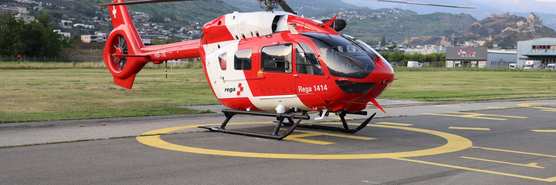 Ein Rega Airbus H145 Rettungshelikopter steht auf einem Landeplatz vor einer Bergkulisse.