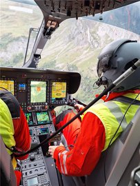 Un pilote de la Rega est assis dans le cockpit de l'hélicoptère. On peut voir les instruments de vol et les commandes. Un paysage alpin avec une cascade est visible en arrière-plan.
