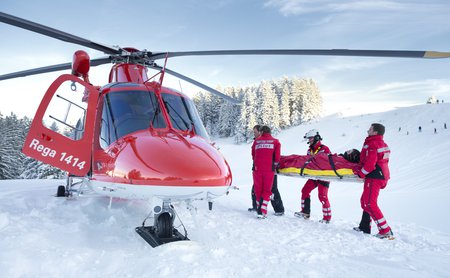 Die Rega-Crew trägt mit Hilfe des Pistenpatrouilleurs einen verletzten Wintersportler zum Helikopter.