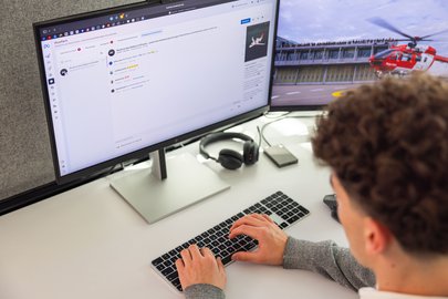 Über die Schulter eines Mediamatiker-Lehrlings geblickt, der am Schreibtisch tippt. Auf einem Monitor ist ein Projektmanagement-Tool, auf dem anderen ein Rega-Helikopter zu sehen.