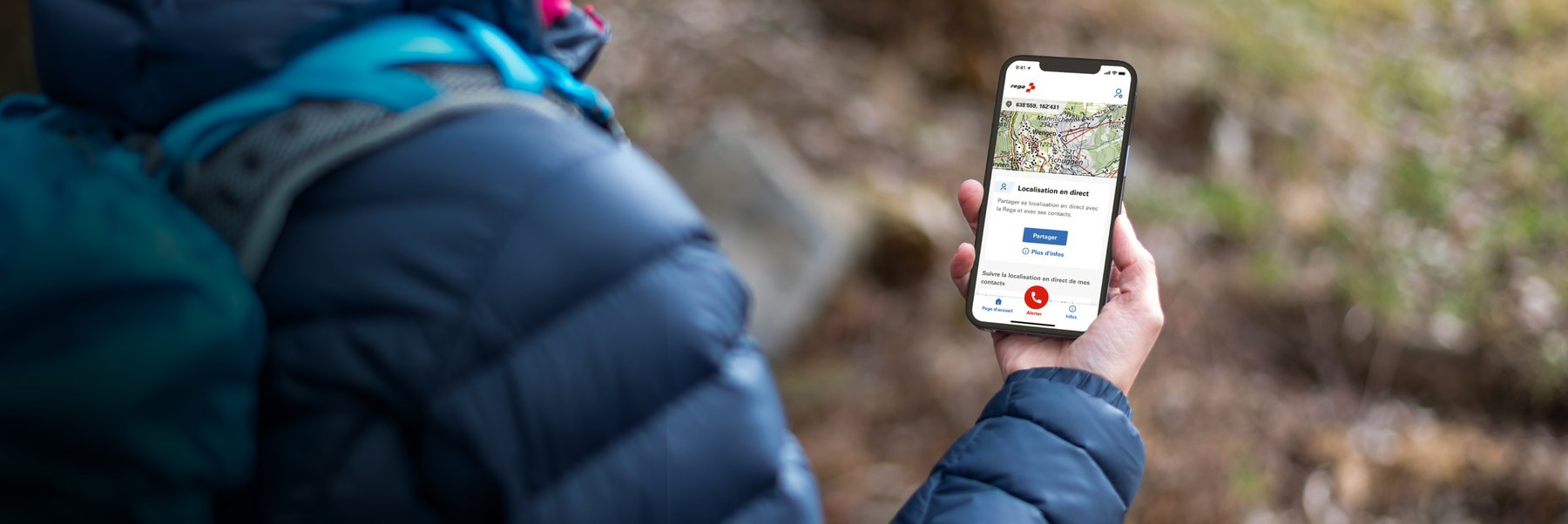 Une personne en extérieur tient un smartphone dans la main, sur lequel l'application Rega est ouverte, affichant la localisation en direct et l'option d'appel d'urgence.