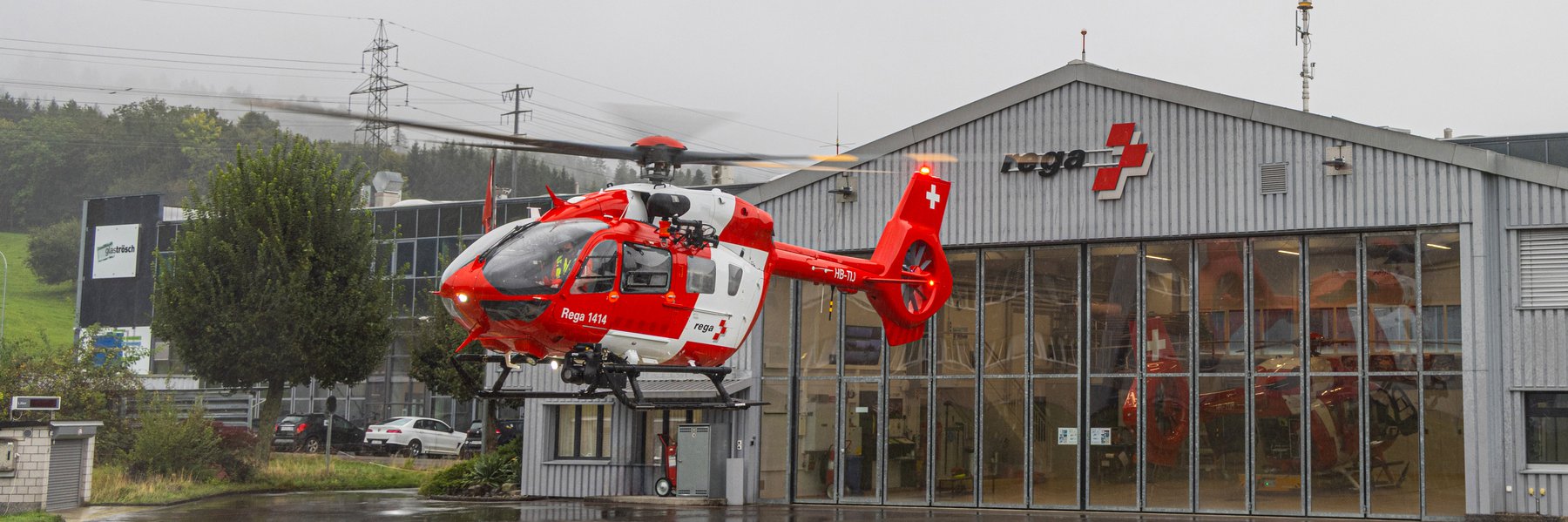 Rettungshelikopter der Schweizer Rega hebt vor einem Hangar bei Regen ab.