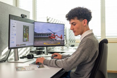 Ein Mediamatiker-Lehrling der Rega sitzt im Büro an einem Schreibtisch mit zwei Monitoren und gestaltet in einem Grafikprogramm ein Layout. Auf dem rechten Monitor ist ein Rega-Helikopter.