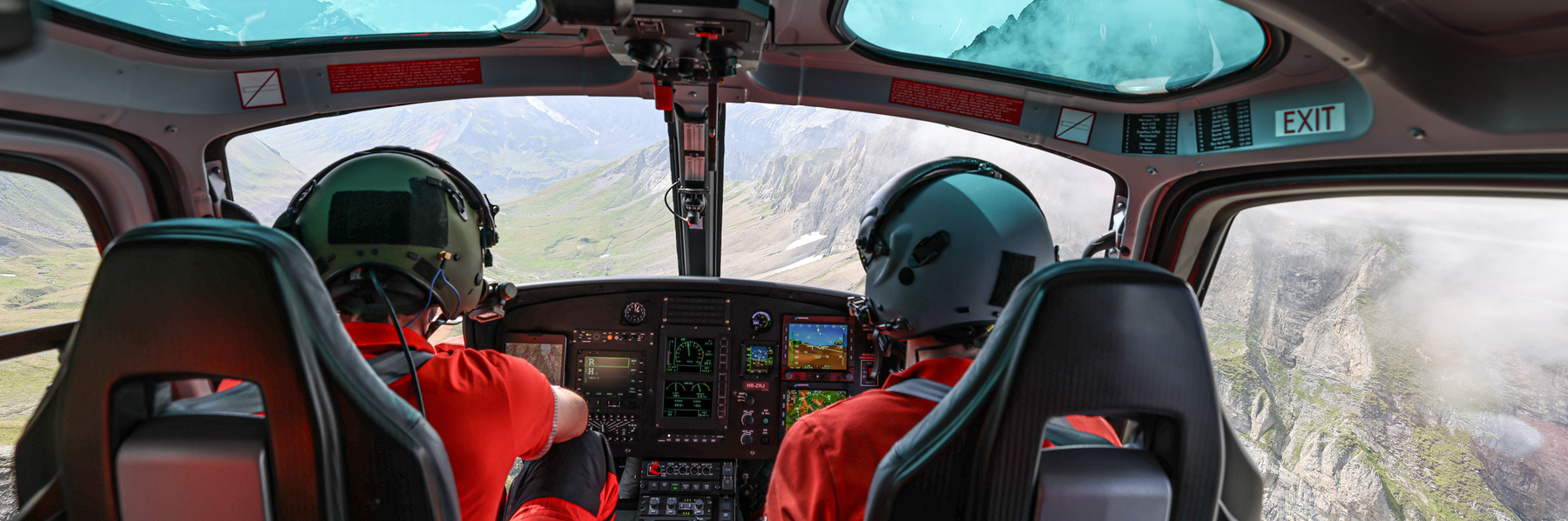 View from behind two Rega pilots in the helicopter cockpit during a flight over a high-alpine, rocky mountain landscape.