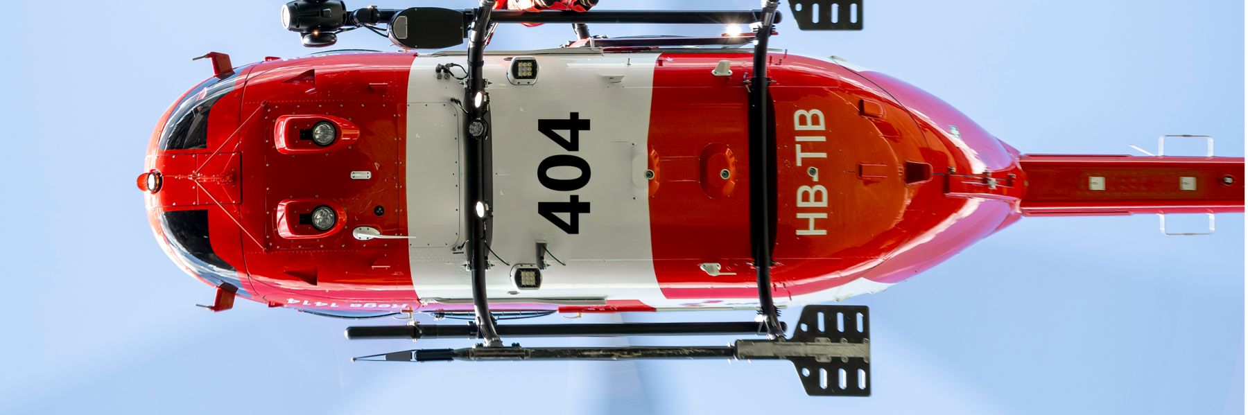 Rega-Helikopter (ein Airbus Helicopters H145, Kennzeichen HB-TIB), von unten vor blauem Himmel aufgenommen. Der rot-weisse Helikopter fliegt im Einsatz mit sichtbarem Hauptrotor.