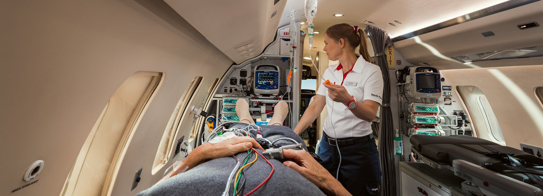  Une employée de la Rega se tient dans un avion-ambulance à côté d'une personne allongée sur une civière et connectée à des équipements médicaux.