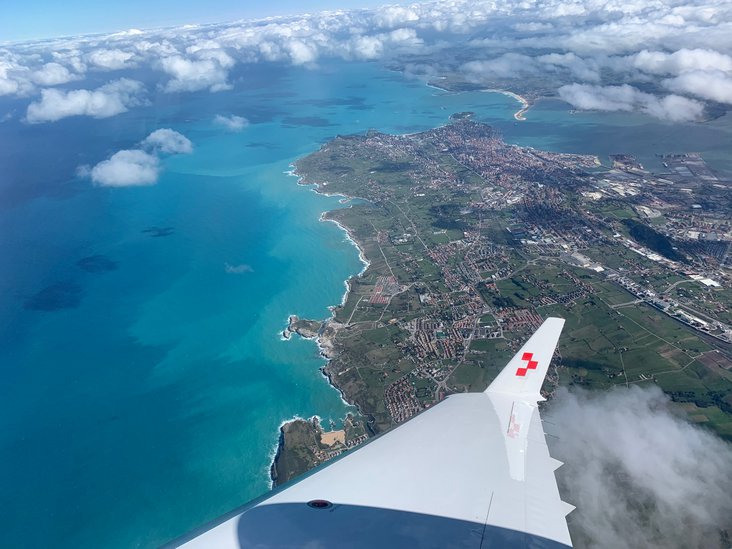 Vista da un jet ambulanza della Rega su una costa con mare turchese, campi verdi e una città sotto uno strato di nuvole.