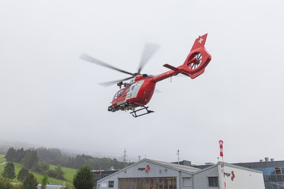Ein roter Rega-Rettungshubschrauber mit weissem Kreuz auf dem Heck fliegt über ein Gebäude mit Rega-Logo. Im Hintergrund sind bewaldete Hügel und ein bewölkter Himmel sichtbar.