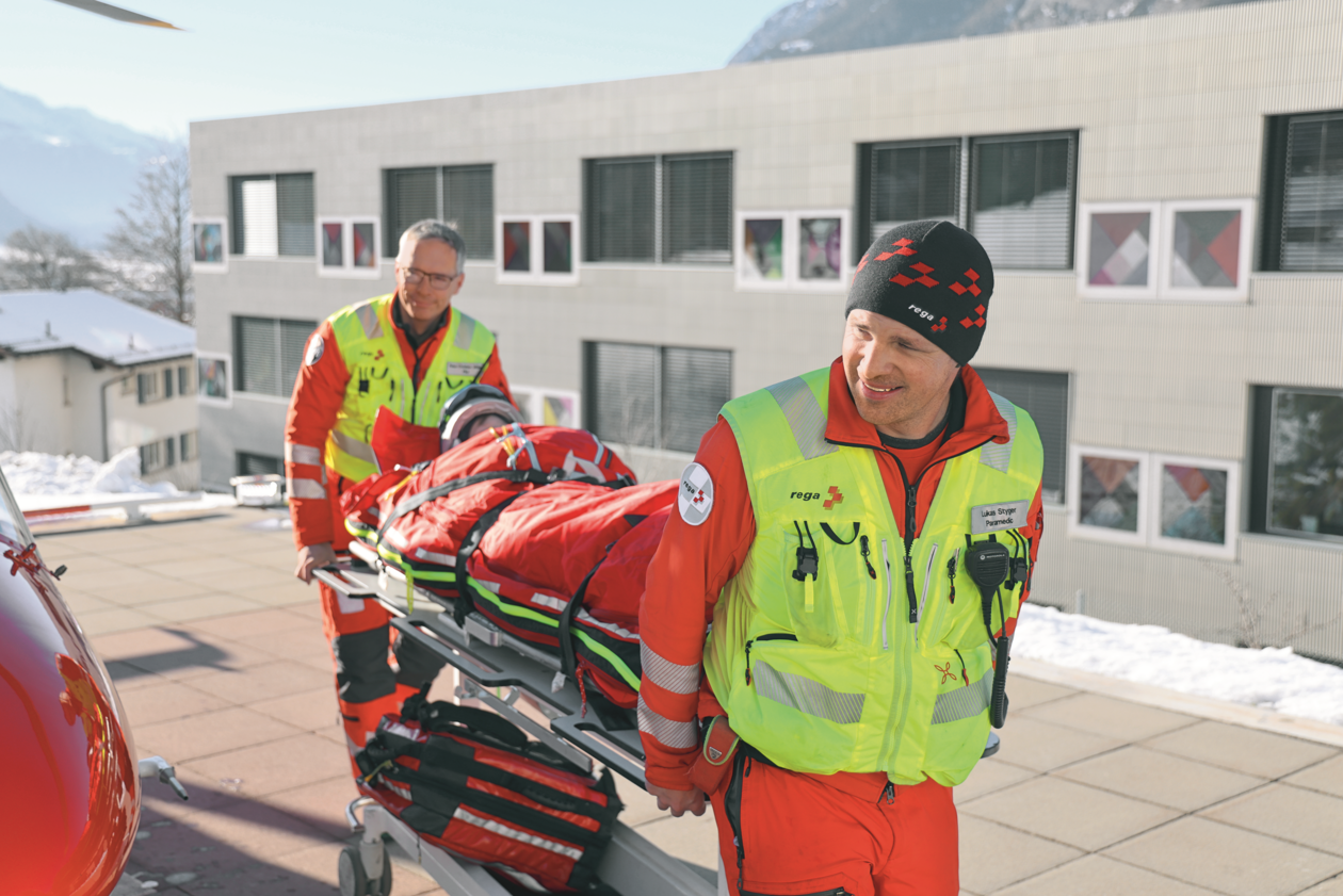 Two Rega crew members in uniform transport a patient on a gurney from the helicopter (partially visible) into a building, presumably a hospital.