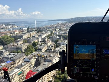 Vue depuis un hélicoptère de sauvetage de la Rega sur la ville de Genève et le lac Léman avec le Jet d'Eau. Au premier plan, des parties du cockpit et des instruments sont visibles.