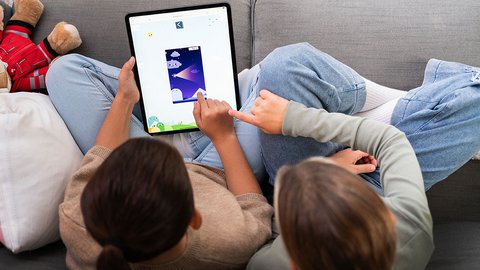 Zwei Kinder liegen auf einem Sofa und spielen gemeinsam ein Spiel auf einem Tablet.