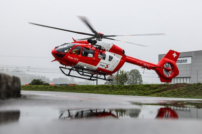 Ein Rega-Rettungshubschrauber mit der Kennung HB-TIJ landet vor einem Gebäude. Der rot-weiße Helikopter spiegelt sich in einer Wasserpfütze auf dem Boden, im Hintergrund ist eine grüne Landschaft sichtbar.