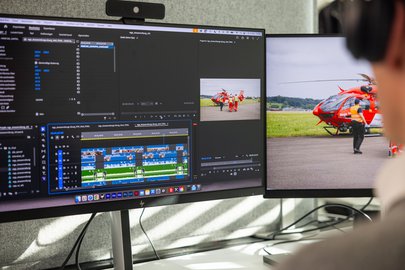Nahaufnahme von zwei Monitoren beim Videoschnitt. Der linke Monitor zeigt die Timeline eines Schnittprogramms, der rechte Monitor die Videovorschau einer Rega-Crew beim Helikopter.