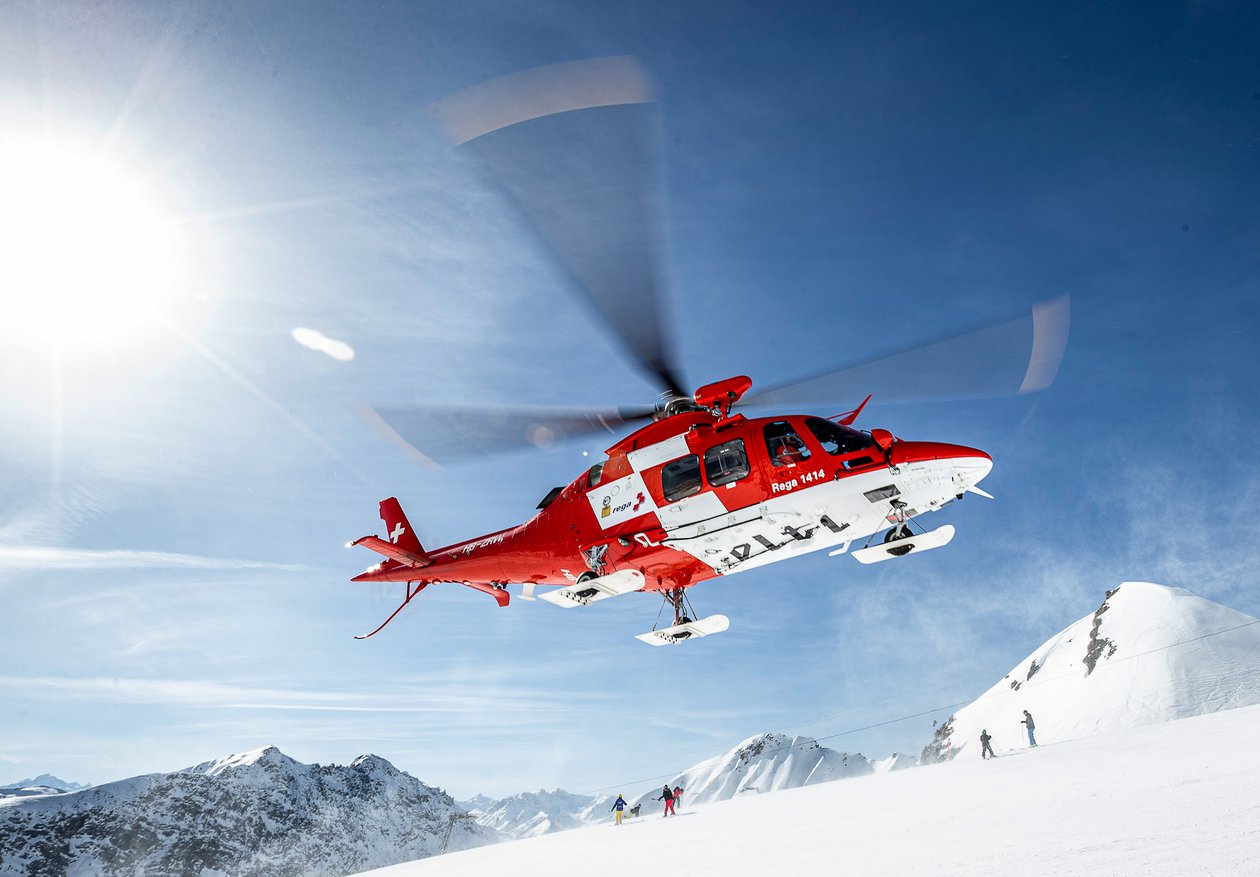 AgustaWestland rescue helicopter against a blue sky and snow-covered mountains