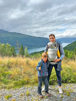Daniela Bergamin, pilota di jet, durante un'escursione con suo figlio nel marsupio e un altro ragazzo in primo piano. Sullo sfondo si vede un paesaggio montano con un lago.