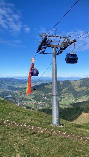 Un parapentiste s'est retrouvé coincé dans les câbles d'une installation de téléphérique dans un paysage montagneux. La voile de parapente, de couleur rouge, jaune et verte, est prise entre un pylône et une télécabine qui passe.