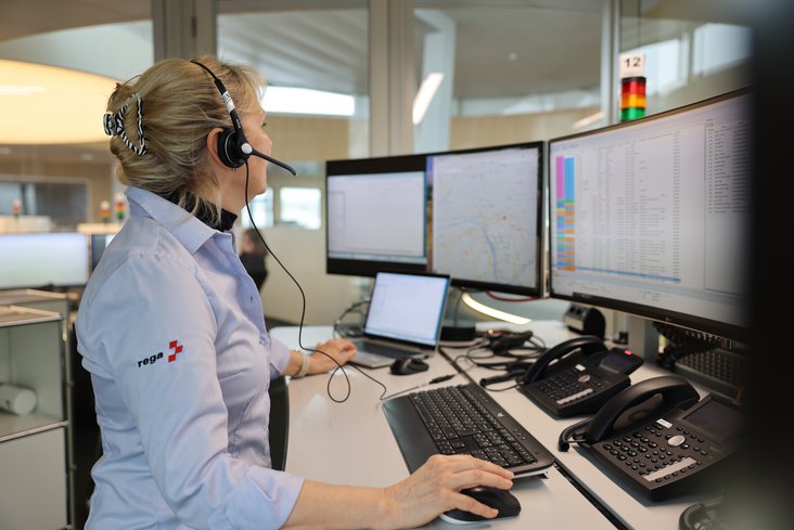 Une médecin-conseil de la Rega portant une chemise blanche et bleue est assise au centre d'opérations devant trois moniteurs d'ordinateur. Elle porte un casque et utilise une souris pour gérer les informations sur les écrans et répondre aux appels d'urgence.