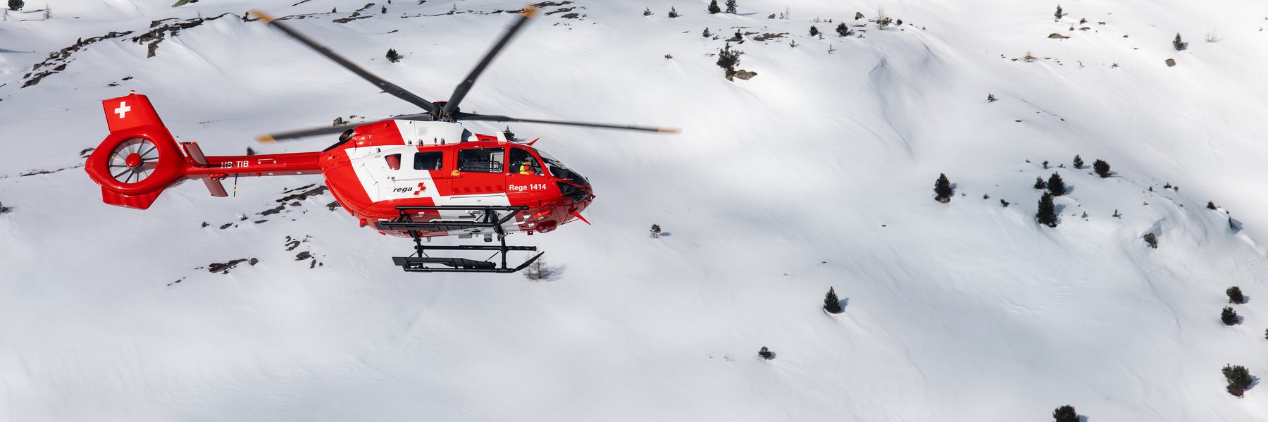 Un elicottero Rega Airbus H145 rosso e bianco, con la sigla HB-TIB, vola sopra un vasto paesaggio montano pesantemente innevato e privo di alberi. L'immagine illustra una missione invernale.