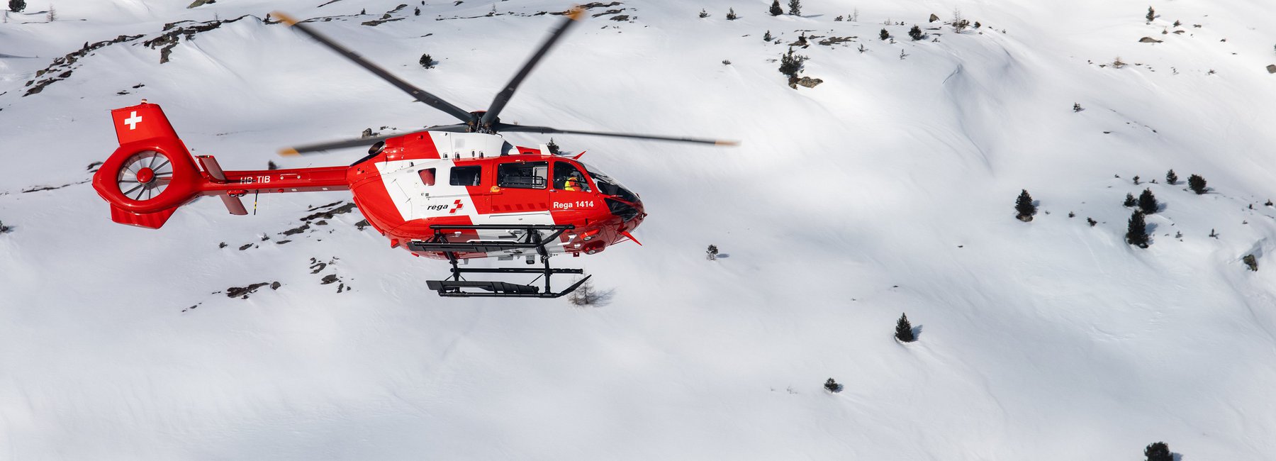 Ein roter und weisser Rega Airbus H145 Helikopter mit der Immatrikulation HB-TIB fliegt über eine weitläufige, tief verschneite und unbewaldete Berglandschaft. Das Bild zeigt den Helikopter bei einem Einsatz im Winter.