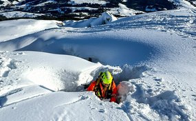 Eine Rettungsperson in orangefarbener Ausrüstung und gelbem Helm steigt aus einer Schneespalte hervor. Hinter ihr liegen weite Berge und ein verschneites Tal.