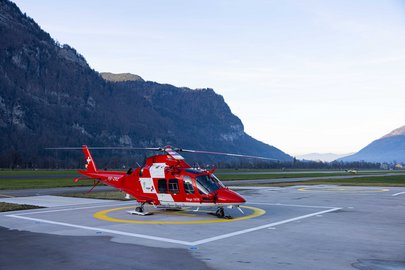 Roter Rega-Helikopter vom Typ AgustaWestland AW169 auf dem Landeplatz der Basis Mollis, umgeben von der Berglandschaft.